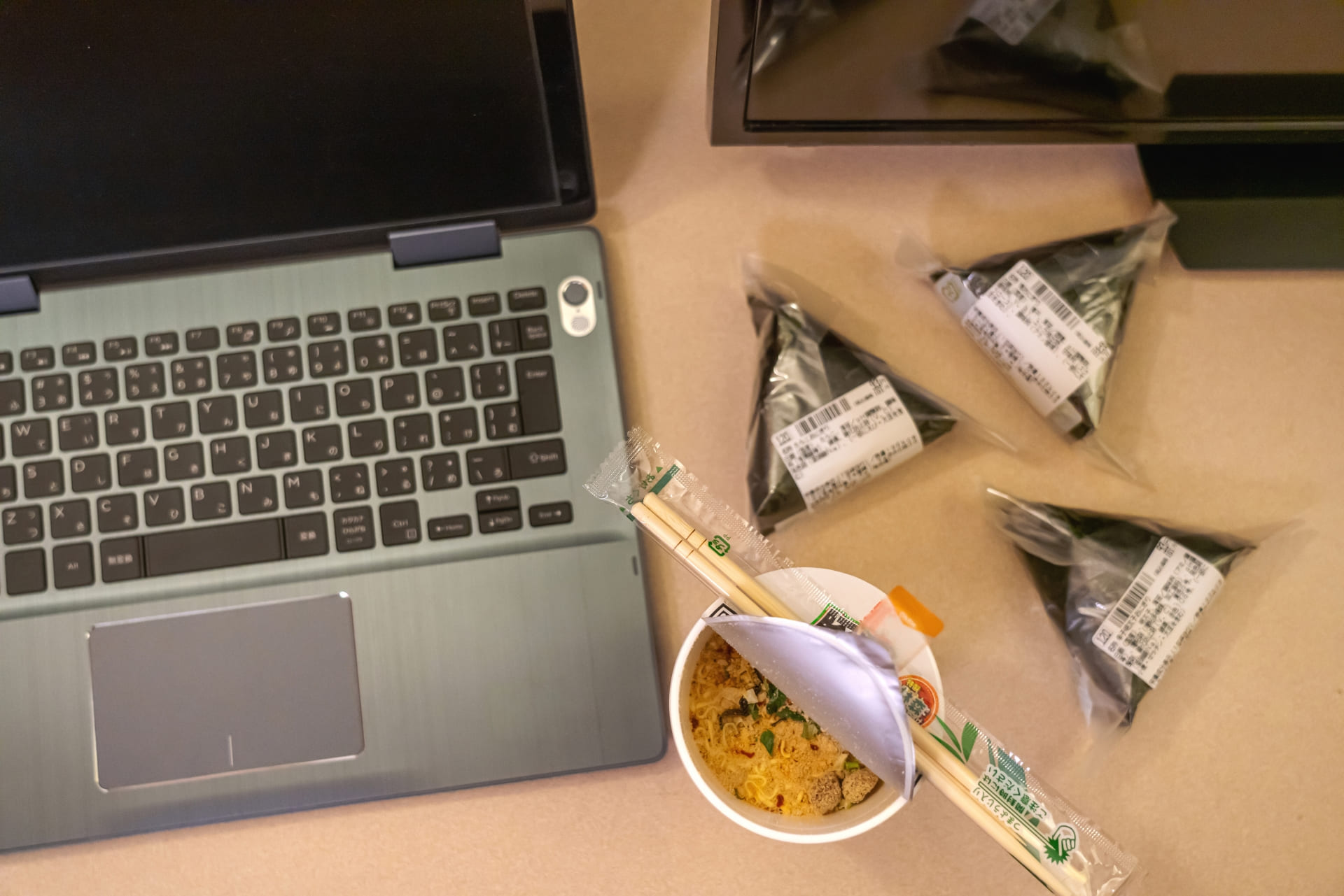 ノートパソコンの横に置かれたカップ麺とおにぎりなどの夜食